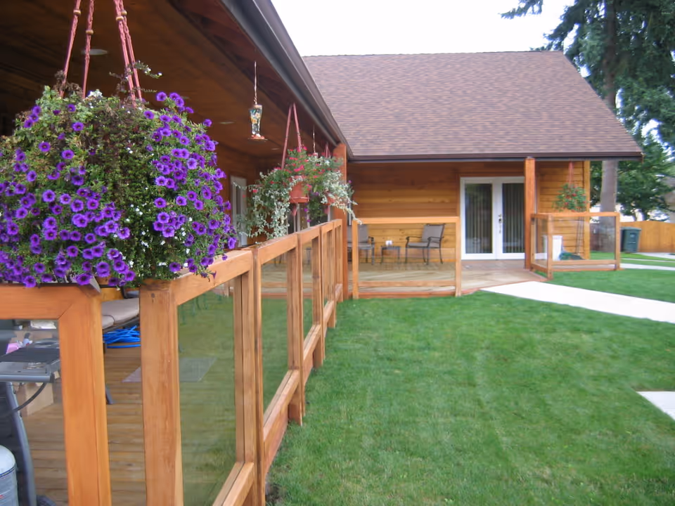 View of a wooden porch with glass panel railings and hanging flower baskets with purple and white flowers. The porch is attached to a wooden building with a sloped roof. There are two chairs and a small table on the porch, and a green lawn with a concrete walkway in front.