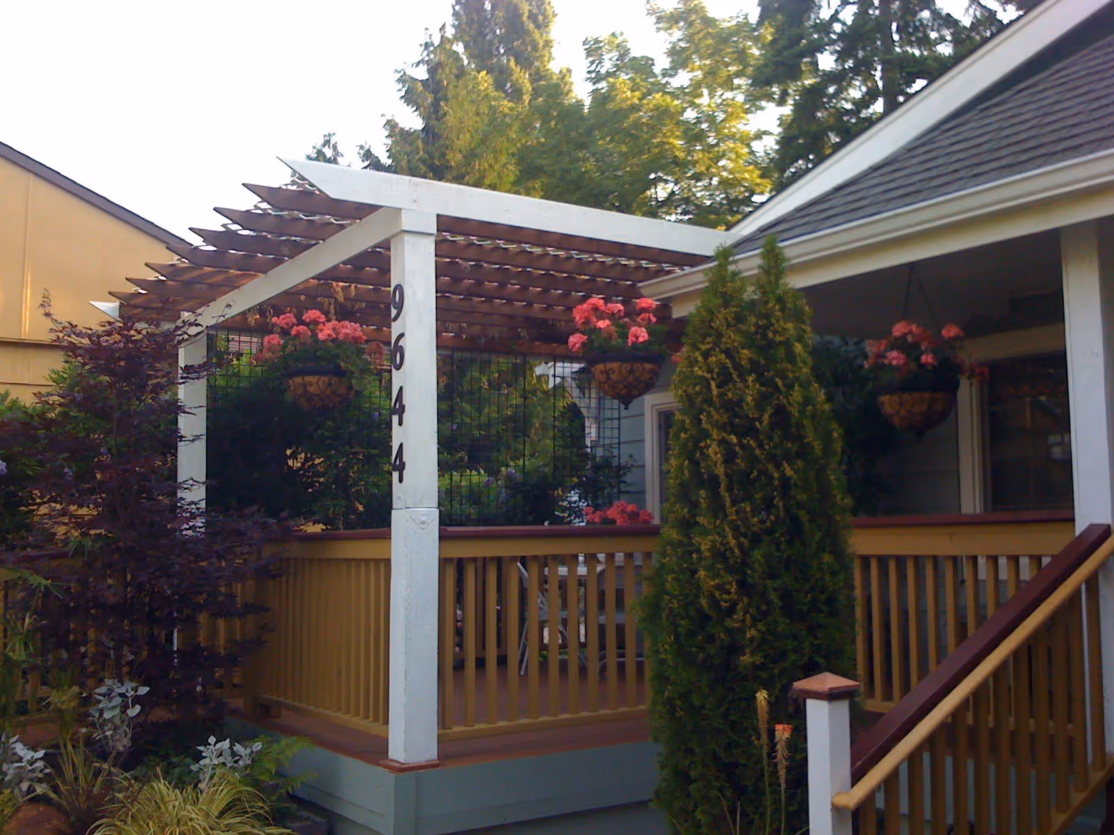 A wooden porch with a pergola overhead, decorated with hanging flower baskets containing pink flowers. The porch has railings and stairs leading up to it. There are various green plants and trees around the porch, and the number 9644 is displayed vertically on one of the pergola posts.