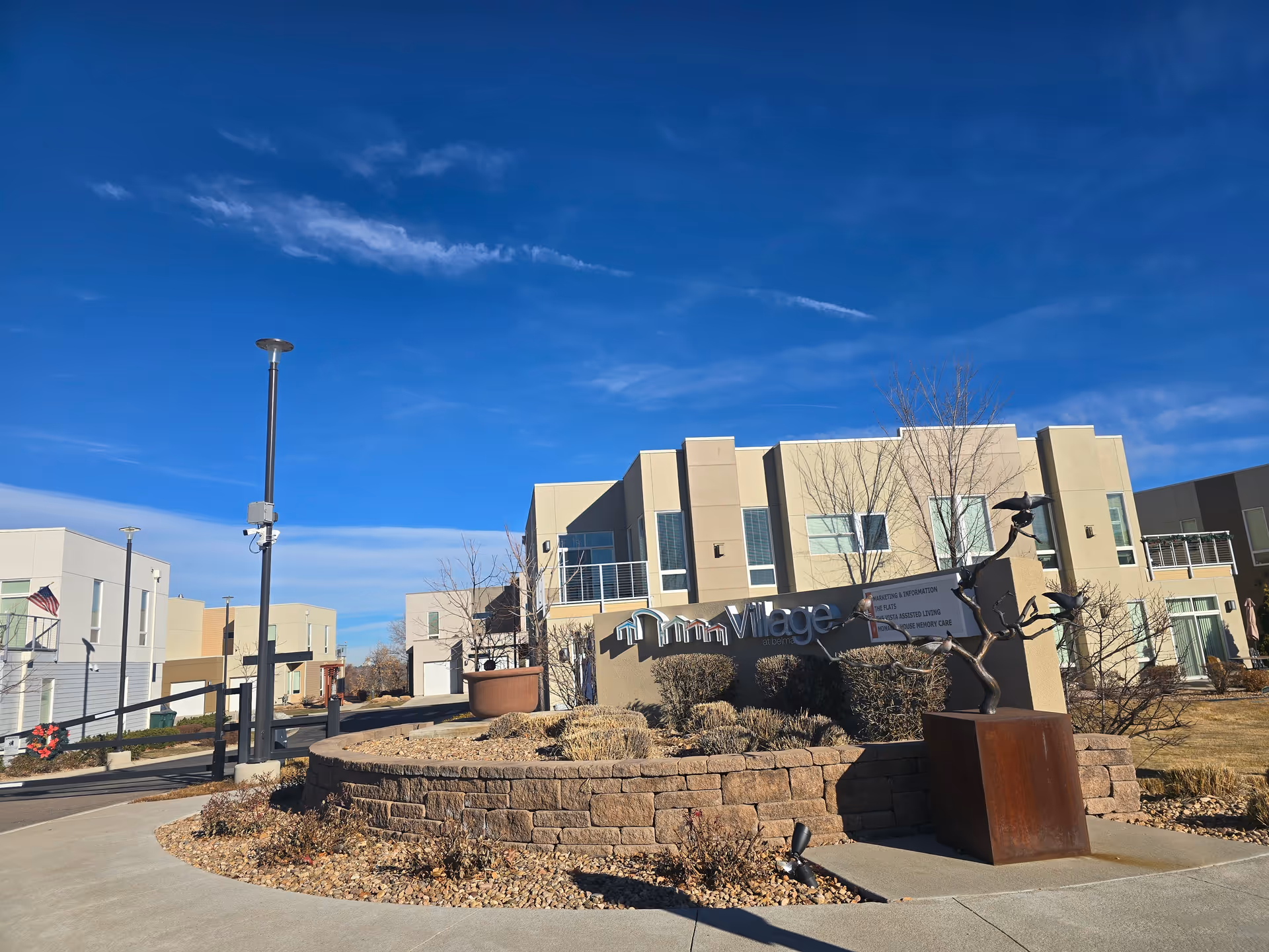 Exterior front of a modern senior living complex with a 'Village' sign, landscaped stone planter, and metal sculpture under a clear blue sky.