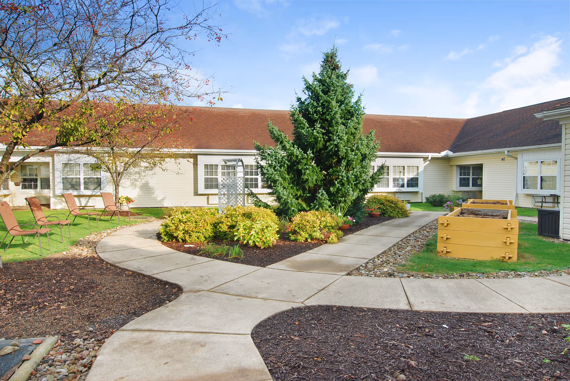 Outdoor courtyard area at a senior living facility with a paved walkway, green grass, bushes, a large evergreen tree, several brown chairs arranged on the lawn, and raised garden beds. The building with beige siding and multiple windows surrounds the courtyard under a clear blue sky.
