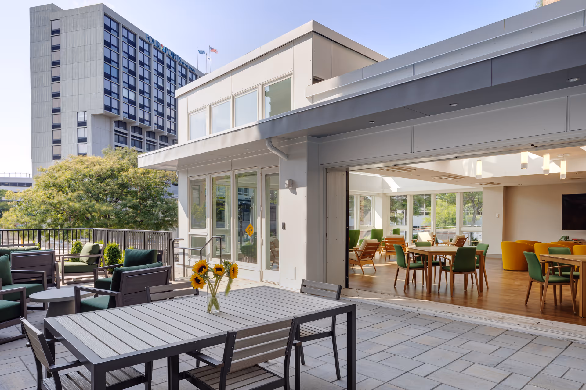 Outdoor patio with tables, chairs and a vase of sunflowers opening into a bright indoor dining/lounge area.