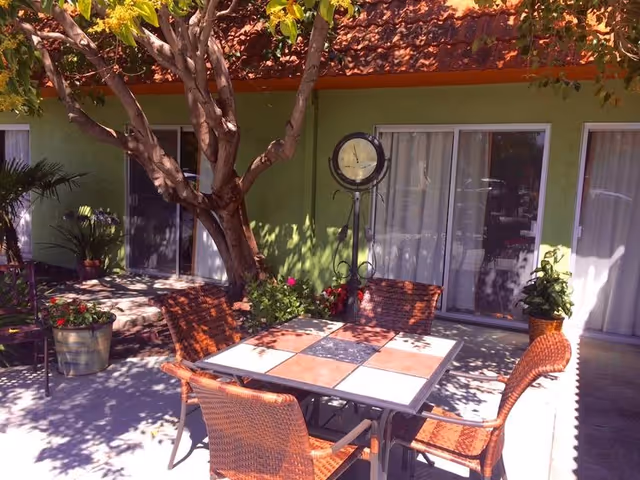 Sunlit outdoor patio with a tiled table and wicker chairs under a tree in front of sliding glass doors on a green building.