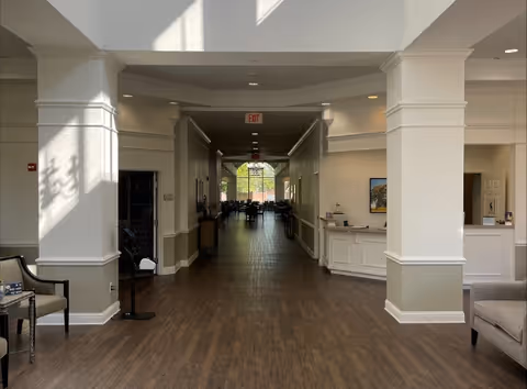 Interior view of a senior living facility hallway with wooden flooring, white walls, and columns. There are chairs on the left and right sides near the entrance, a reception desk on the right, and a dining area visible at the end of the hallway with large windows letting in natural light.