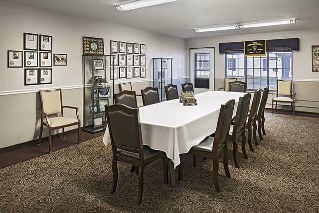 A formal dining room with a long rectangular table covered with a white tablecloth, surrounded by wooden chairs with cushioned seats. The room has patterned carpet flooring, framed pictures and certificates on the walls, glass display cabinets, and large windows with a door leading outside.