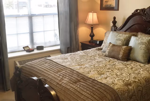 Sunlit bedroom featuring a carved wooden bed with patterned bedding, a bedside table and lamp next to a window.