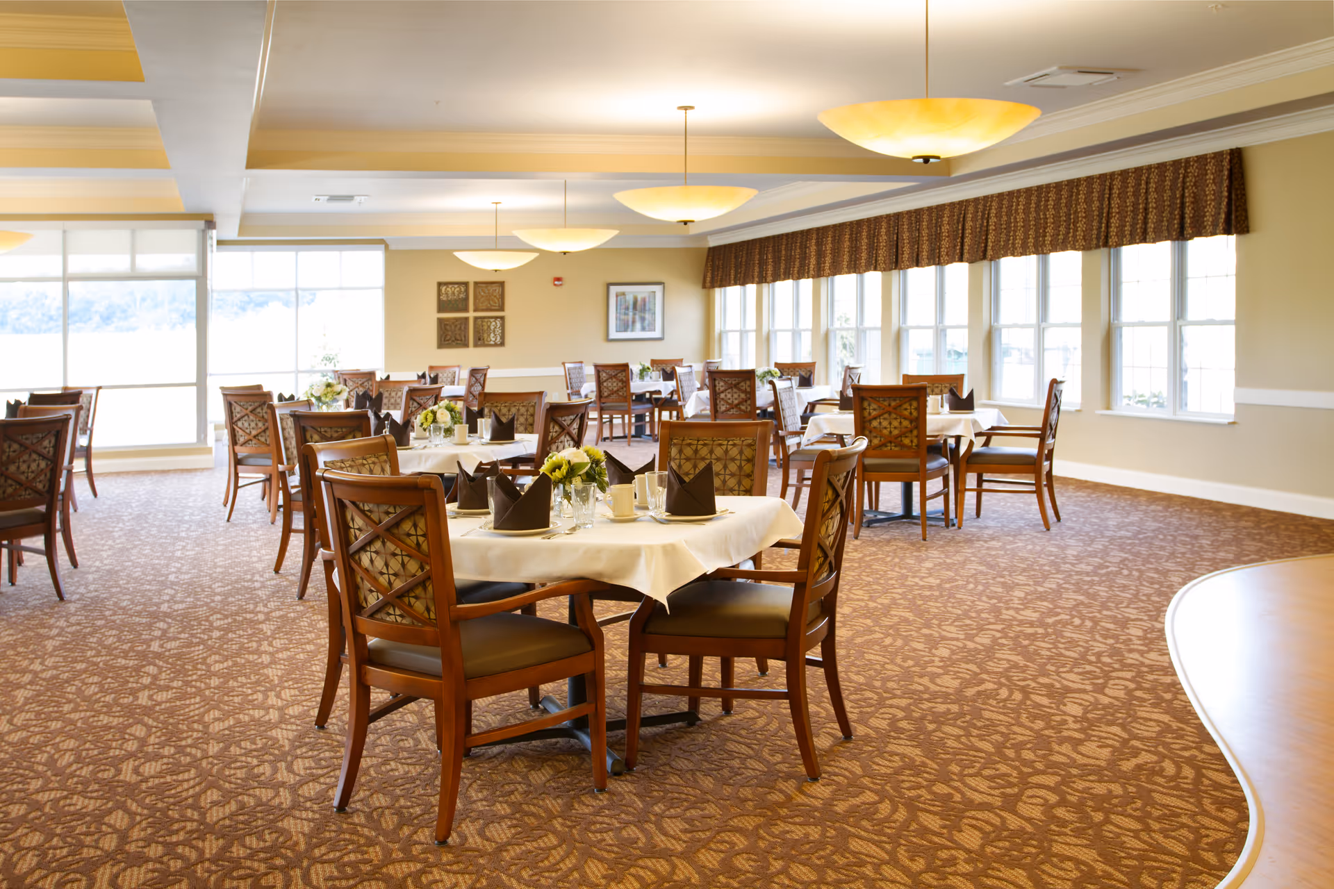 A bright dining room with multiple round tables covered with white tablecloths, each set with folded dark napkins, glassware, and floral centerpieces. The room has large windows along one wall letting in natural light, patterned carpet, and warm ceiling lights.