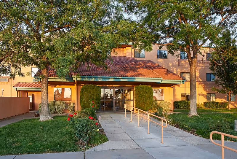 Front exterior view of Sandia Vista Senior Living building with a walkway leading to glass entrance doors, surrounded by green grass, trees, and bushes.
