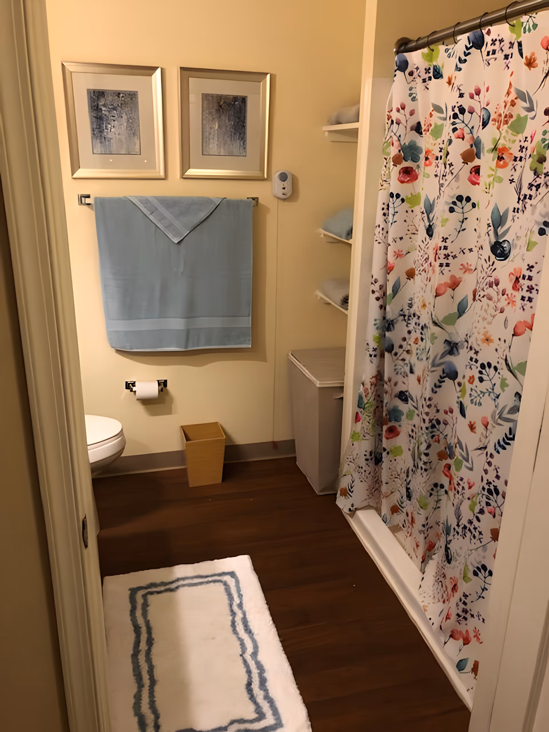A bathroom with a floral shower curtain, a white toilet, a blue towel hanging on a towel rack, two framed pictures on the wall above the towel, a small wooden trash bin, a white and blue bath mat on a wooden floor, and shelves with folded towels.