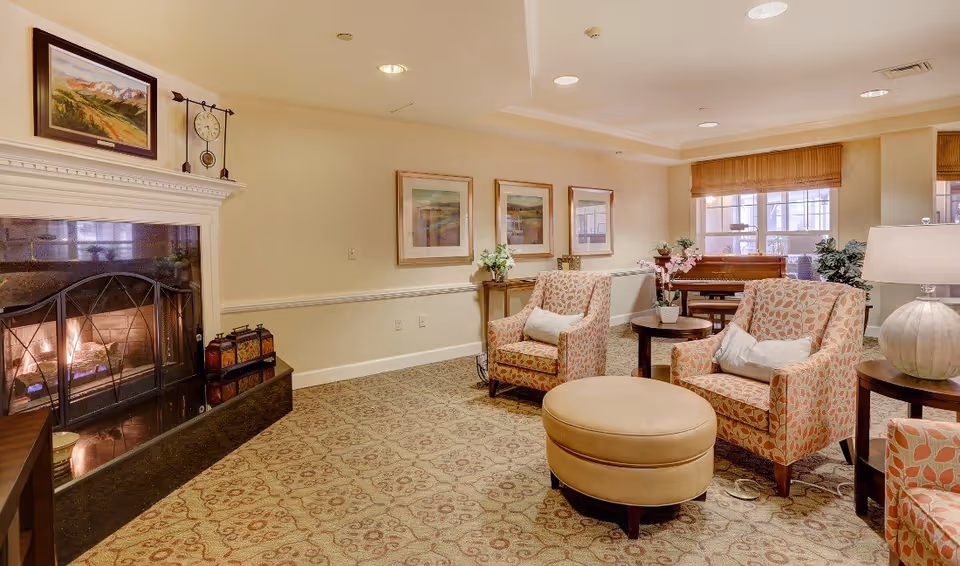 A cozy living room area in a senior living facility with a lit fireplace on the left, two patterned armchairs with white cushions, a round beige ottoman, side tables with lamps and plants, framed artwork on the walls, and large windows with wooden blinds letting in natural light.