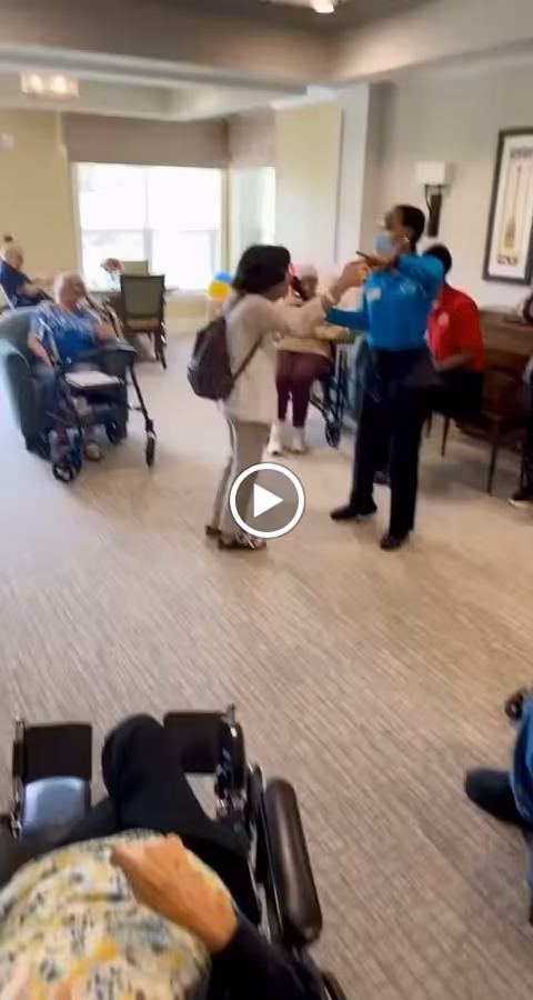 A group of elderly people in a senior living facility common area. Some are seated in wheelchairs and chairs, while a staff member wearing a mask interacts with a visitor. The room has large windows letting in natural light and framed artwork on the walls.
