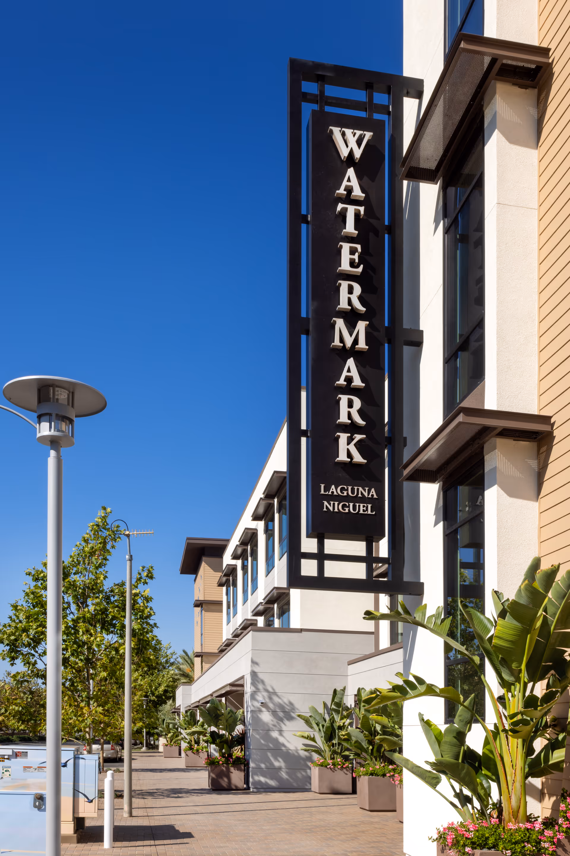 Exterior view of the Watermark Laguna Niguel building with a large vertical sign displaying the name. The building is modern with large windows and surrounded by plants and trees under a clear blue sky.