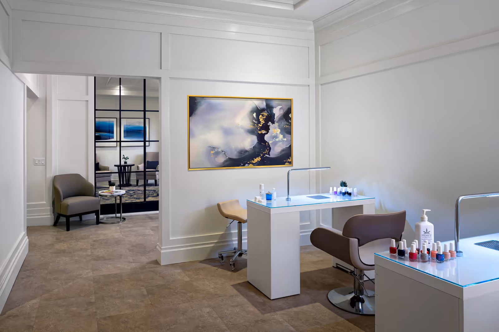 Manicure salon area with two white nail tables, brown salon chairs, nail polish bottles, and seating visible through a glass partition.