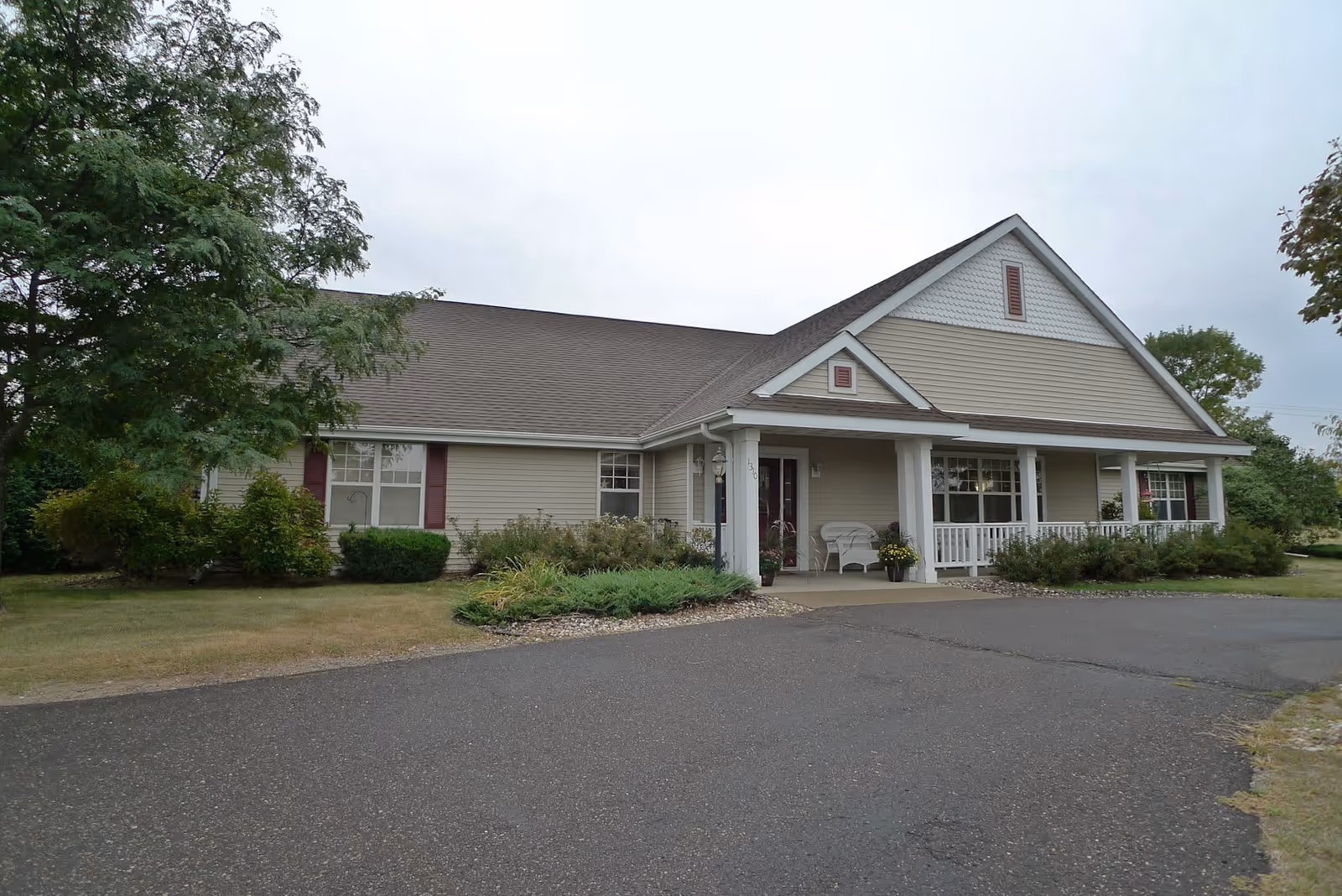Exterior view of a single-story senior living facility building with beige siding, a gabled roof, and a covered front porch with white columns. The building is surrounded by green shrubs, trees, and a paved driveway in front.