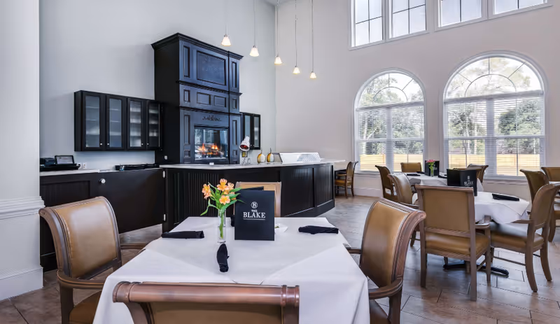 A bright dining room with large arched windows letting in natural light. The room features several tables covered with white tablecloths, each set with black napkins and a small vase of flowers. There is a dark wooden fireplace and cabinetry along one wall, and the chairs are upholstered in light brown leather.