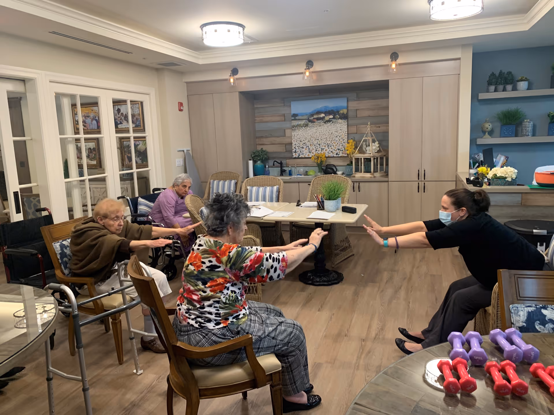A group of elderly women and a caregiver wearing a mask are seated in a room participating in a seated exercise session. The women are stretching their arms forward while sitting on chairs and using walkers. The room has wooden flooring, a table with papers and plants, and shelves with decorative items in the background.