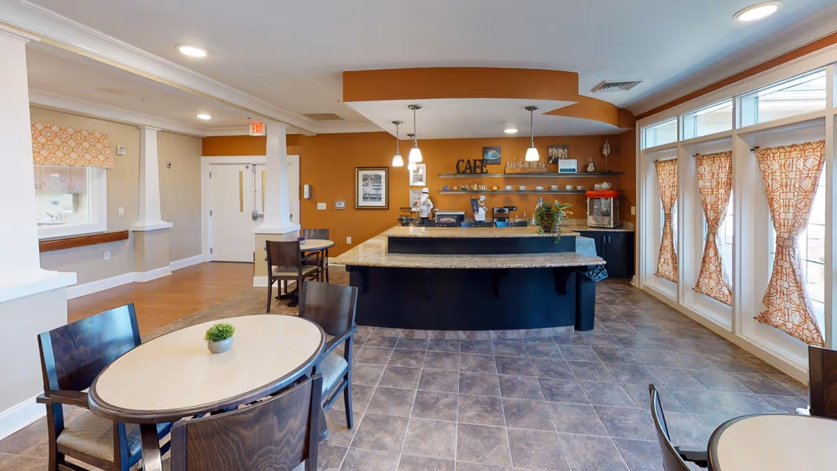 Bright café-style dining area with round tables, chairs, a service counter labeled 'CAFÉ' and large windows with patterned curtains.
