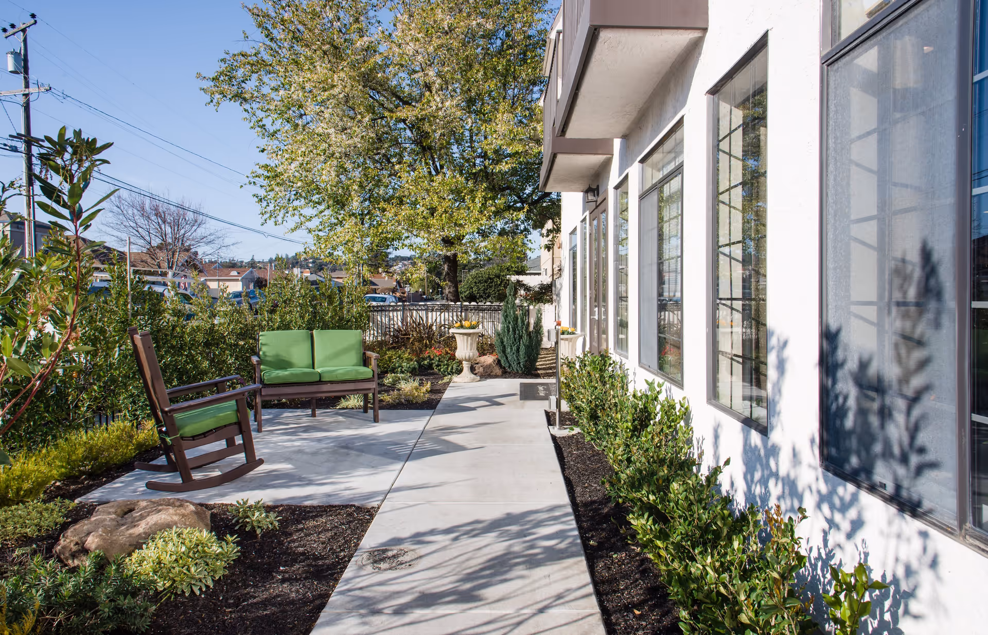 Outdoor patio area at Carefield Castro Valley Assisted Living and Memory Care featuring a concrete walkway, green cushioned wooden rocking chair and loveseat, surrounded by plants and trees with a white building on the right side.