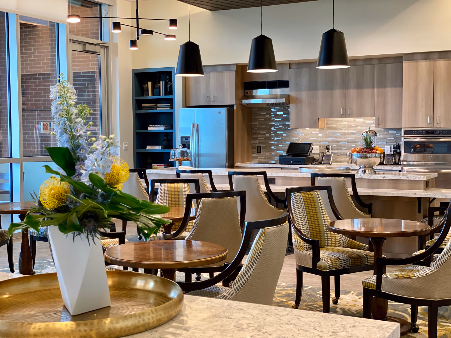 Communal dining area featuring round wooden tables and upholstered chairs, a floral centerpiece in the foreground, and an open kitchen with pendant lights and stainless appliances.