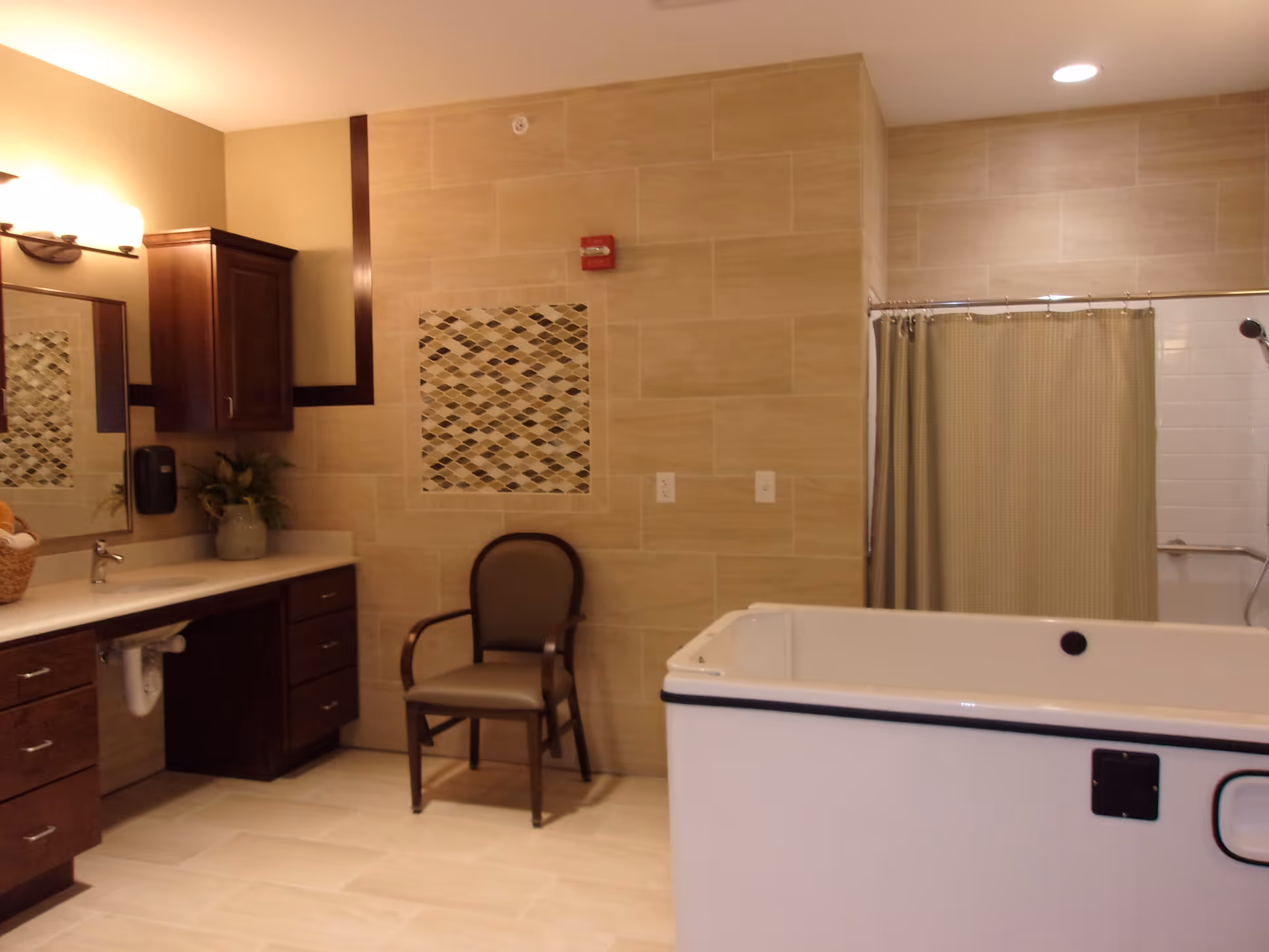 A spacious bathroom with beige tiled walls and floor, featuring a large white bathtub with a black handle, a shower area with a beige curtain, a wooden chair with armrests, a countertop with a sink, dark wood cabinets, a mirror, and a wall-mounted light fixture.