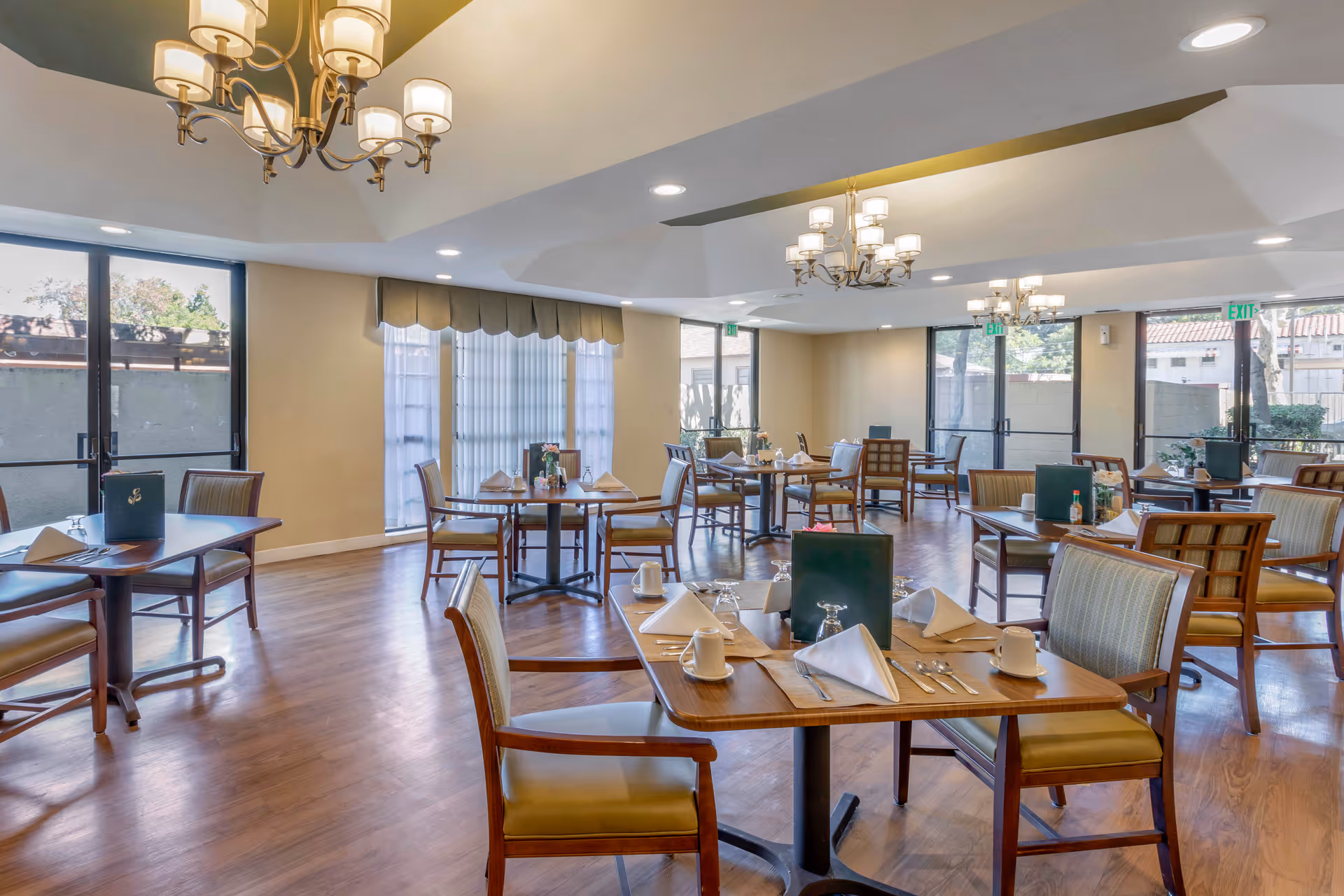 A bright dining room with multiple wooden tables and chairs arranged neatly. Each table is set with white napkins, cups, glasses, and silverware. Large windows and glass doors allow natural light to fill the room. Elegant chandeliers hang from the ceiling, and the floor is a polished wood finish.