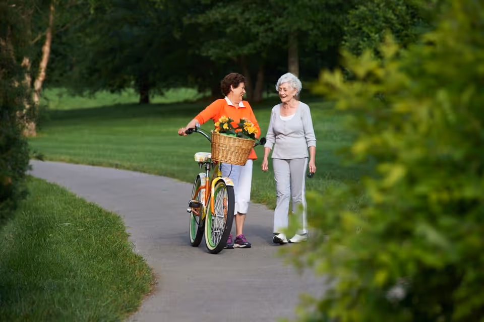 Two elderly women walking together on a paved path in a green park. One woman is pushing a bicycle with a basket of flowers, and both are smiling and engaged in conversation.