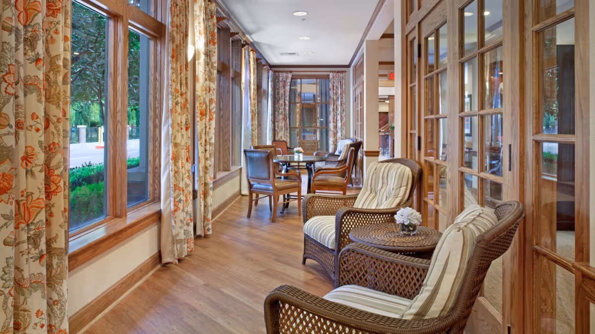 A bright and cozy sitting area with wicker chairs featuring striped cushions and small round tables with flower vases. The space has large windows with floral curtains allowing natural light to fill the room, and wooden framed glass doors along one side. The floor is wooden, and additional chairs and tables are visible further down the hallway.