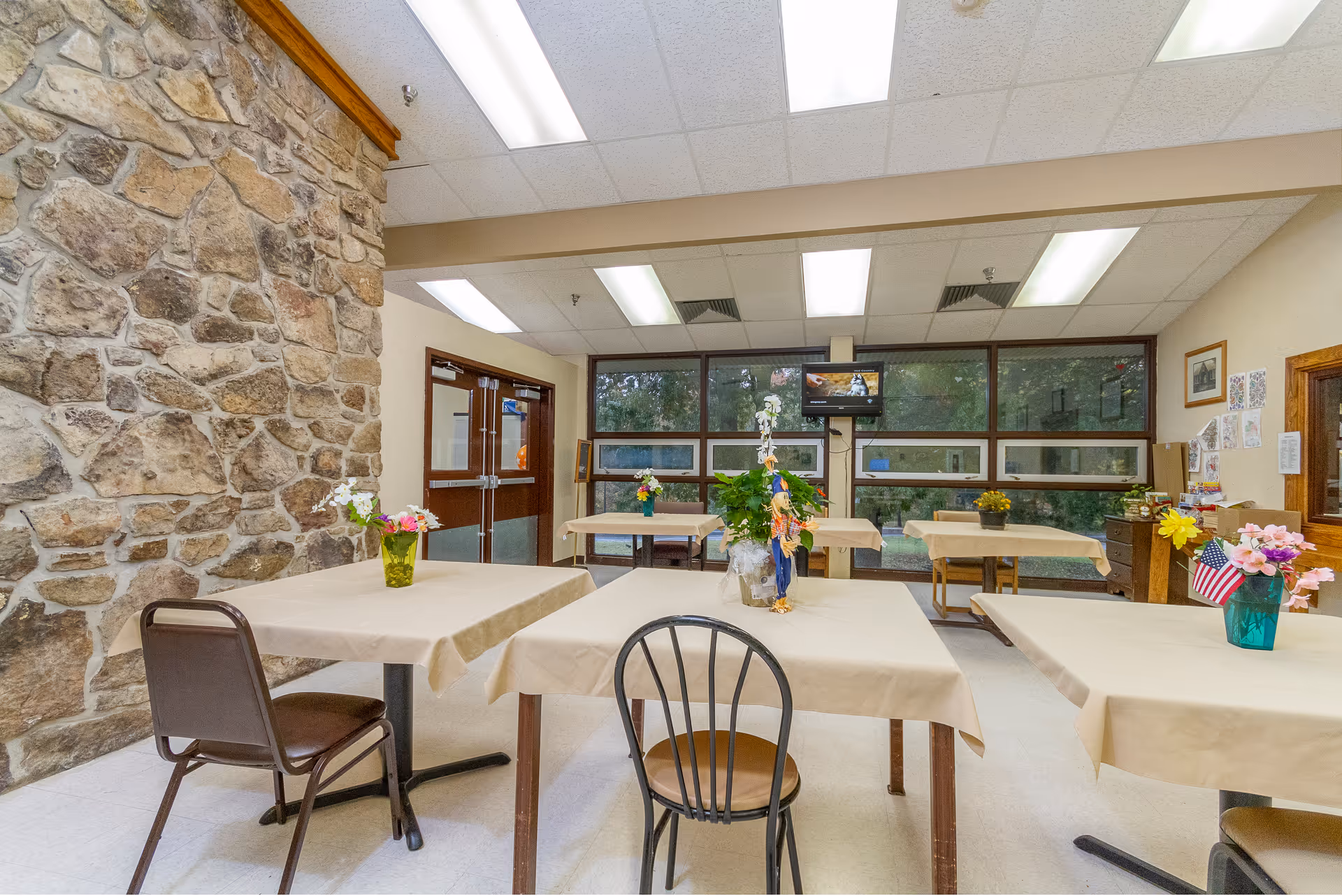 A dining area with several tables covered in beige tablecloths, each decorated with small flower arrangements. The room features a stone wall on the left, large windows on the far side letting in natural light, and a television mounted on the wall. Chairs are placed around the tables, and the space has a clean, simple, and welcoming atmosphere.
