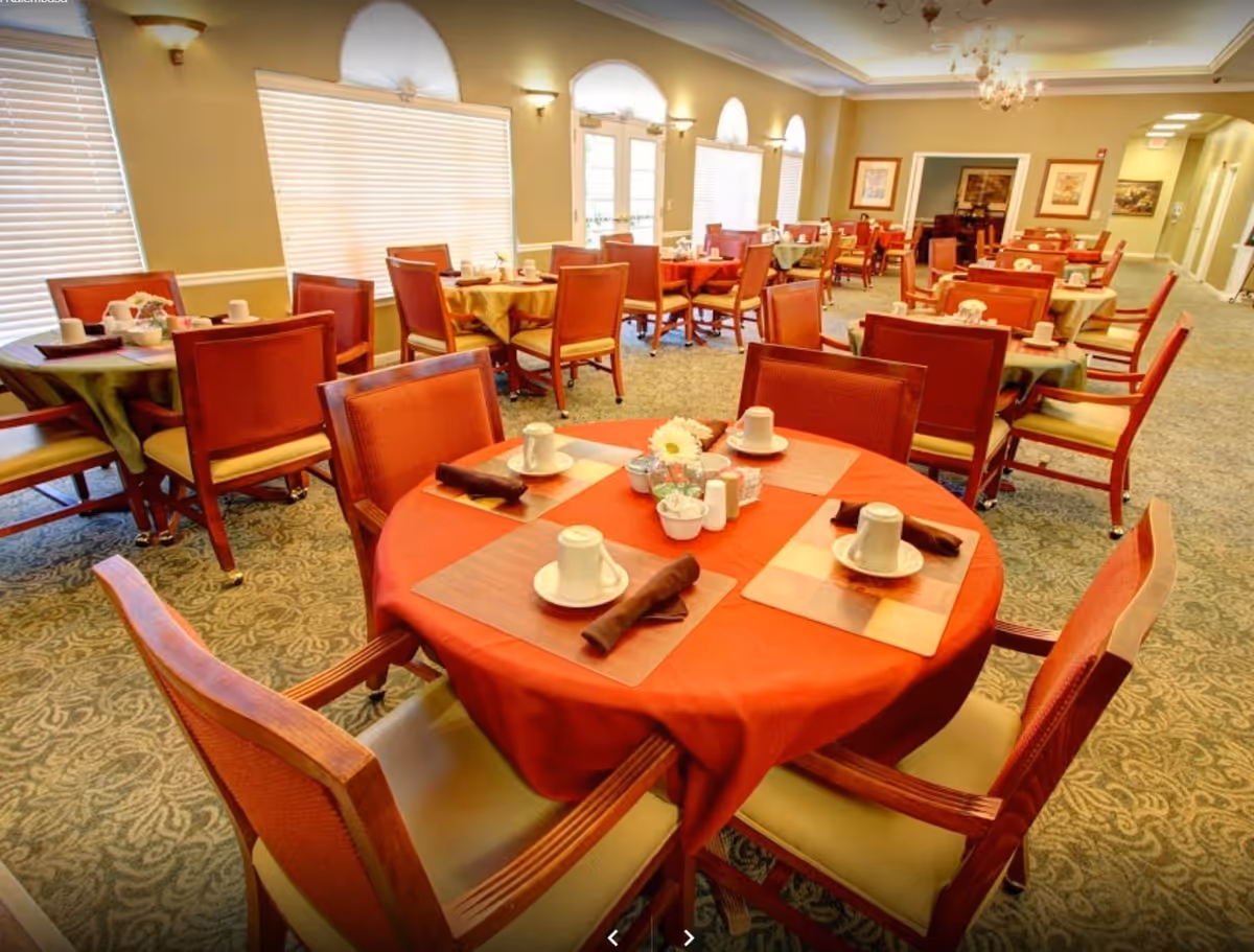 A dining room in a senior living facility with multiple round tables covered with tablecloths, each set with cups, saucers, napkins, and small flower arrangements. The room has large windows with blinds, carpeted floors, and warm lighting.
