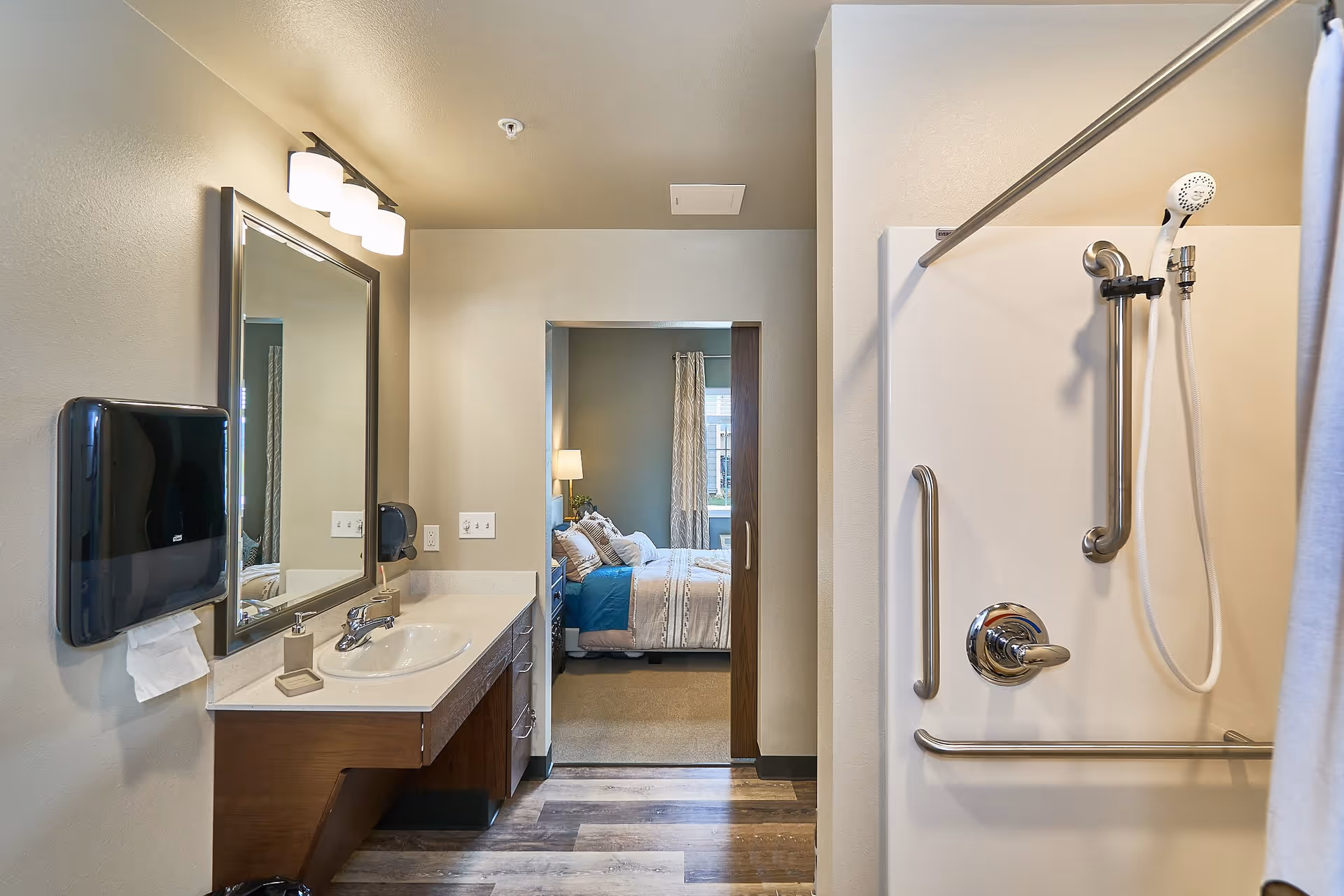 View of a bathroom with a sink, mirror, and paper towel dispenser on the left, and a shower with grab bars and a handheld showerhead on the right. Through an open doorway, a bedroom with a bed, pillows, a lamp, and a window with curtains is visible.