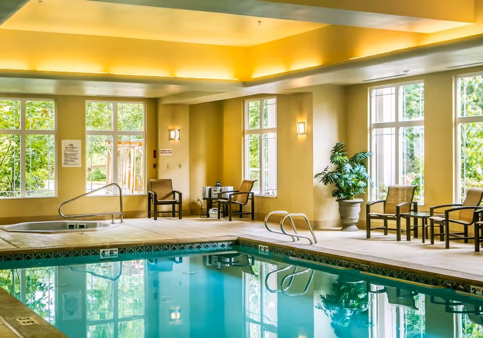Indoor swimming pool area with clear water, surrounded by beige tiled floor and several chairs and tables along the walls. Large windows let in natural light and show greenery outside. The ceiling has recessed lighting creating a warm ambiance.
