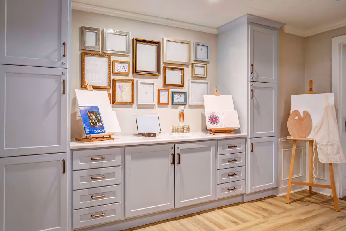 Bright interior art and craft area with white cabinets, countertops, easels, and a gallery wall of empty picture frames.
