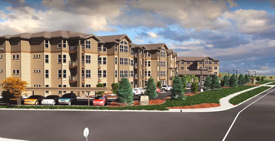 Front exterior of a multi-story senior living building with a parking lot, trees, and landscaped grounds under a cloudy sky.