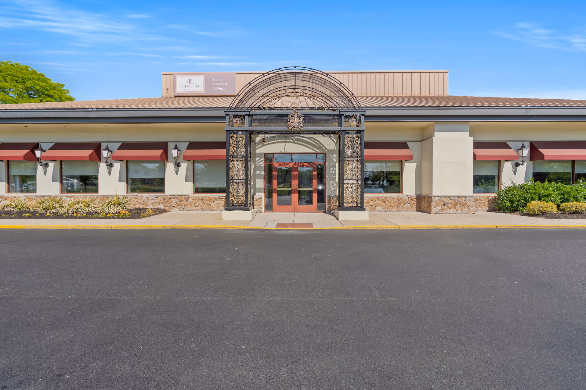 Front entrance of a single-story senior living facility with a decorative metal archway, double glass doors, and red window awnings.