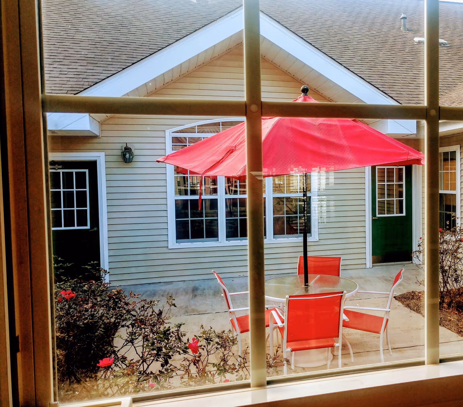 View through a window of a small courtyard patio with a round glass table, red chairs and a red umbrella in front of a light-colored building.
