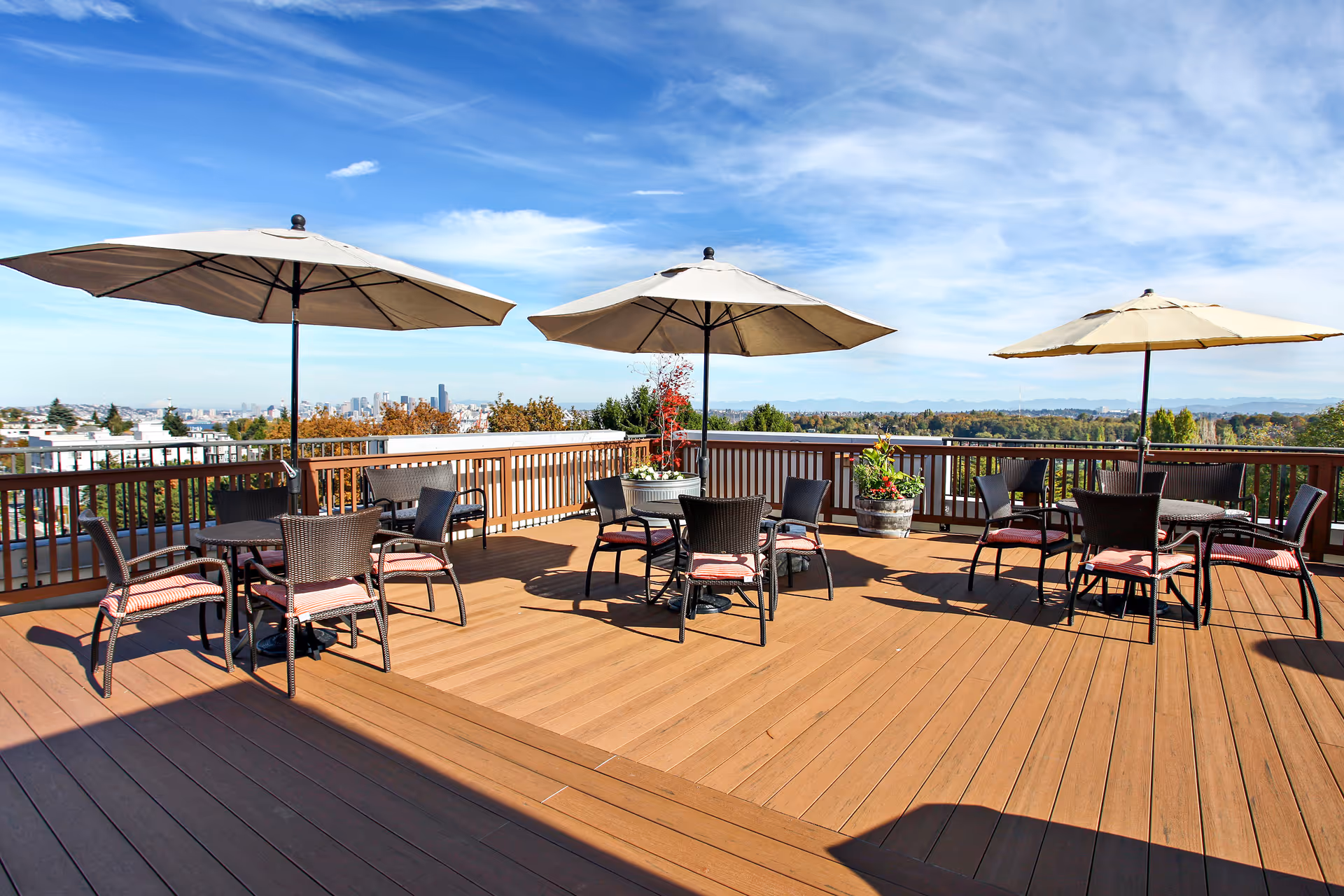 Outdoor patio area with wooden deck featuring three round tables, each with four wicker chairs with red cushions and large beige umbrellas providing shade. The patio overlooks a scenic view of a city skyline, trees, and distant mountains under a blue sky with wispy clouds.