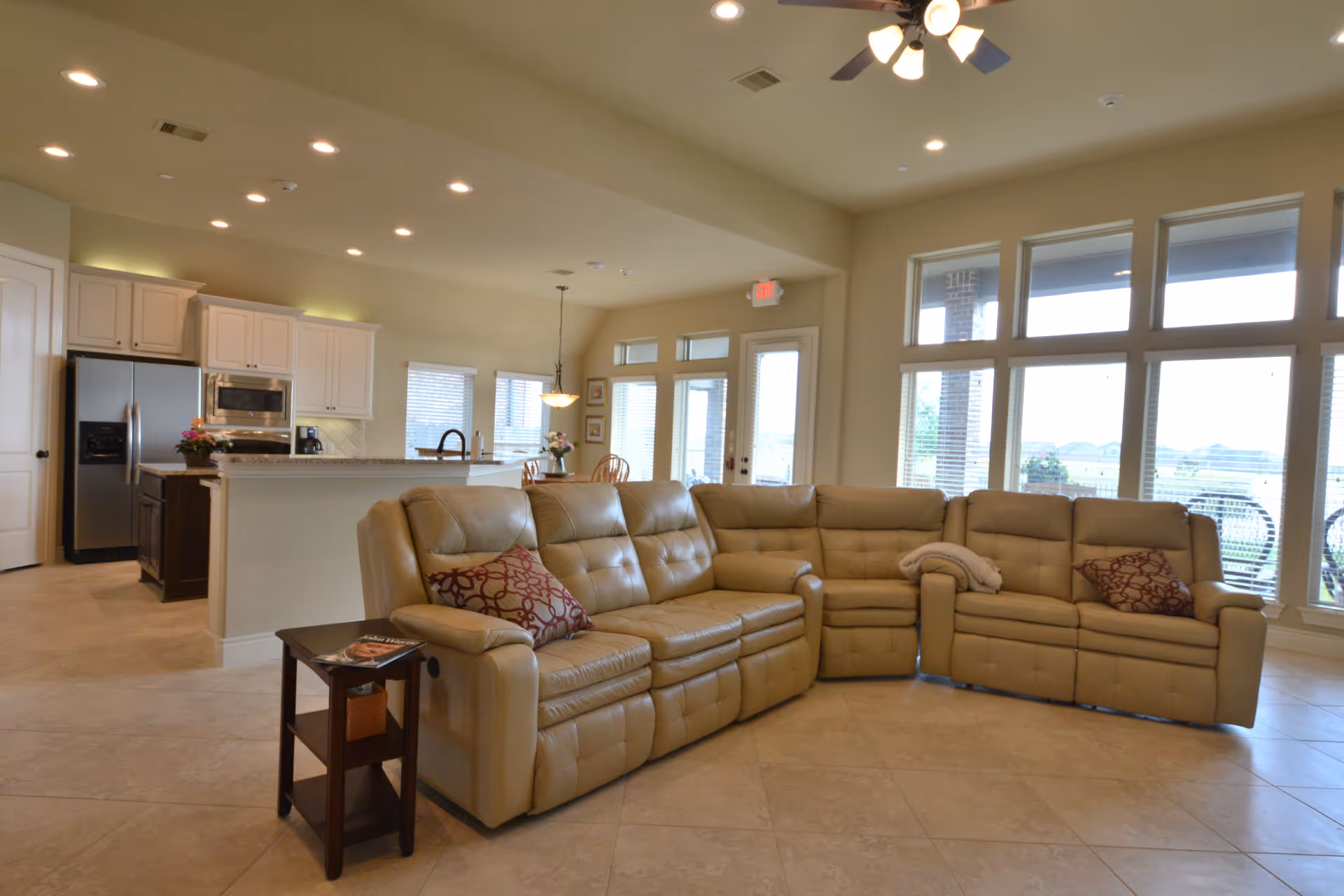 Spacious open living area with a large beige sectional sofa adorned with two patterned pillows and a folded blanket. The room features large windows with blinds allowing natural light to fill the space. In the background, there is a kitchen with white cabinets, stainless steel appliances, and a dining area with a wooden table and chairs. The ceiling has recessed lighting and a ceiling fan with lights.