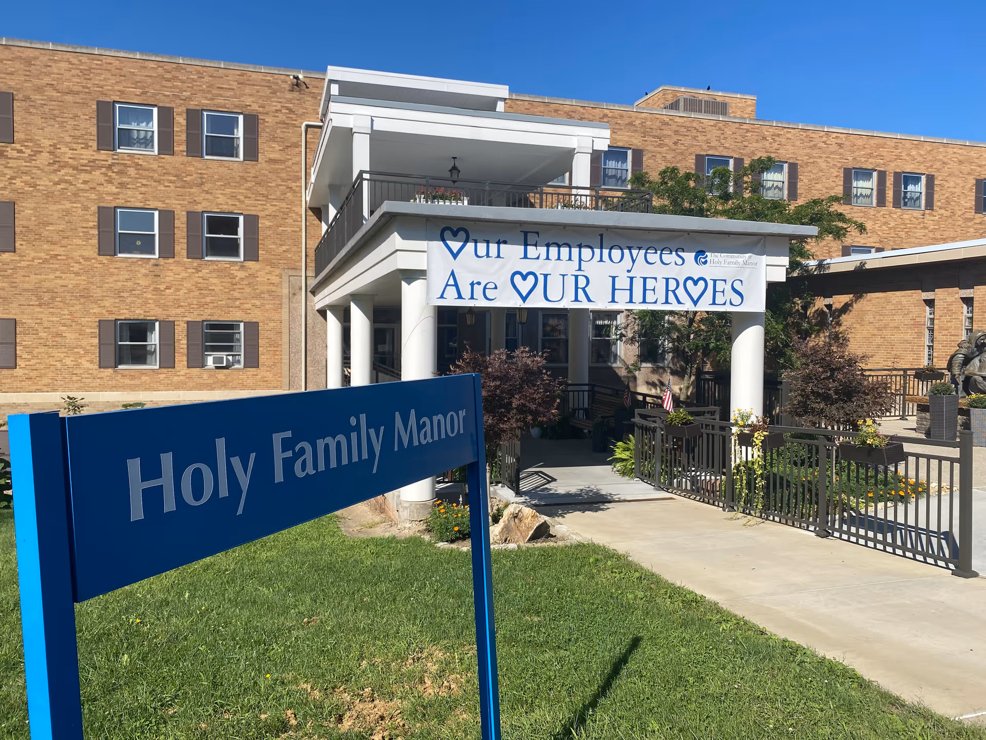 Exterior view of The Community at Holy Family Manor building with a blue sign in the foreground that reads 'Holy Family Manor'. The building is made of brick with multiple windows and a covered entrance with white columns. A banner hanging above the entrance reads 'Our Employees Are Our Heroes' with heart symbols.