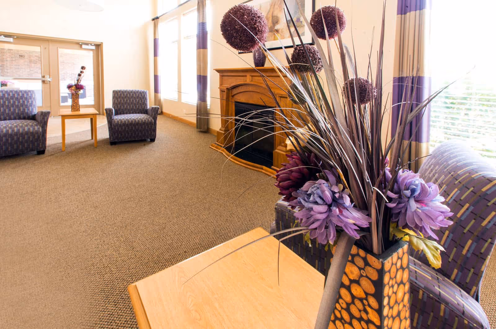 A cozy seating area in a healthcare facility with two patterned armchairs, a wooden table with a decorative vase of purple flowers and dried plants, a wooden fireplace, large windows with purple and beige curtains, and a glass door in the background.