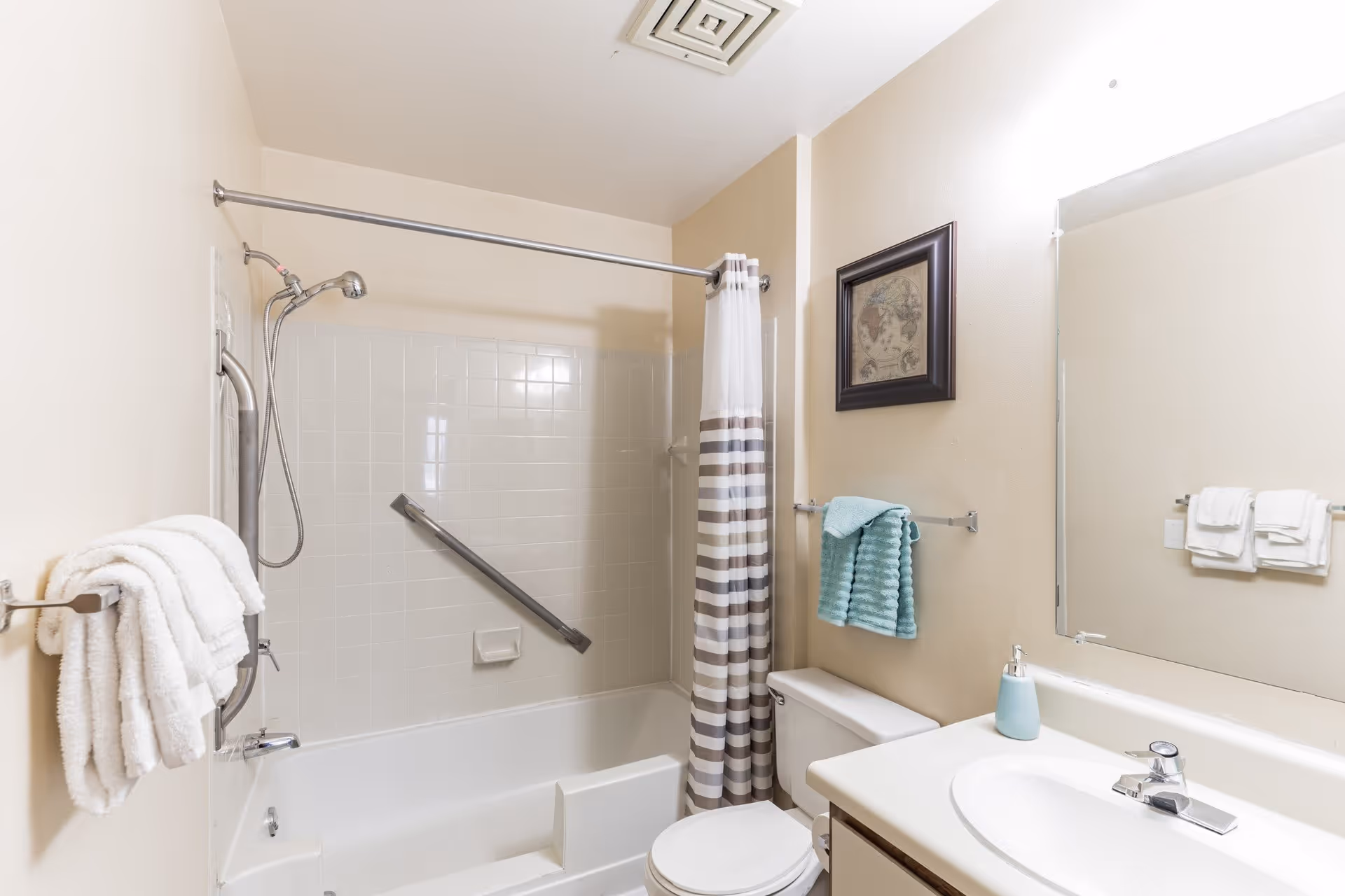 A clean bathroom featuring a bathtub with a showerhead and a grab bar, a striped shower curtain, a toilet, a sink with a soap dispenser, and several towels hanging on racks. There is a framed picture on the wall and a large mirror above the sink.