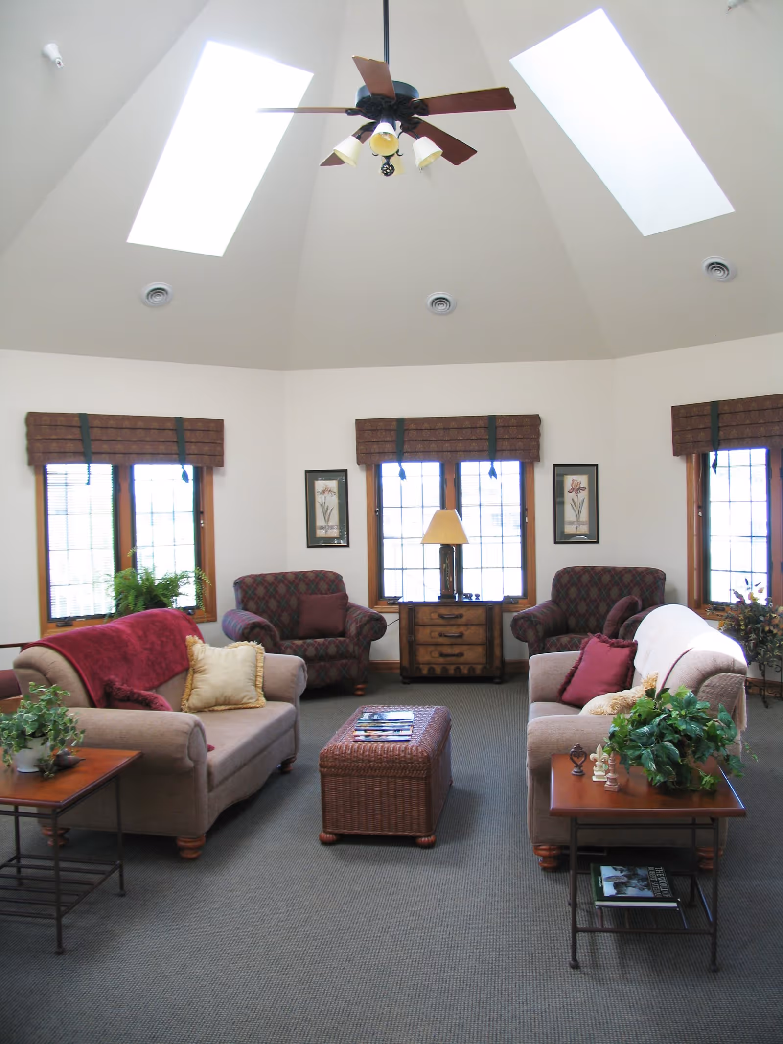 A bright living room with high vaulted ceilings featuring two skylights and a ceiling fan. The room has three windows with brown valances, two patterned armchairs, two beige sofas with red and beige cushions, a wicker coffee table, two wooden side tables with plants, and a wooden cabinet with a lamp between the windows.
