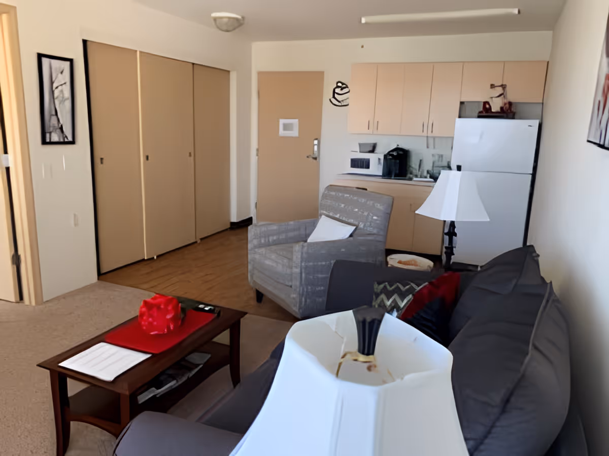 Small living area with a couch, armchair, coffee table and a kitchenette with cabinets and a refrigerator.