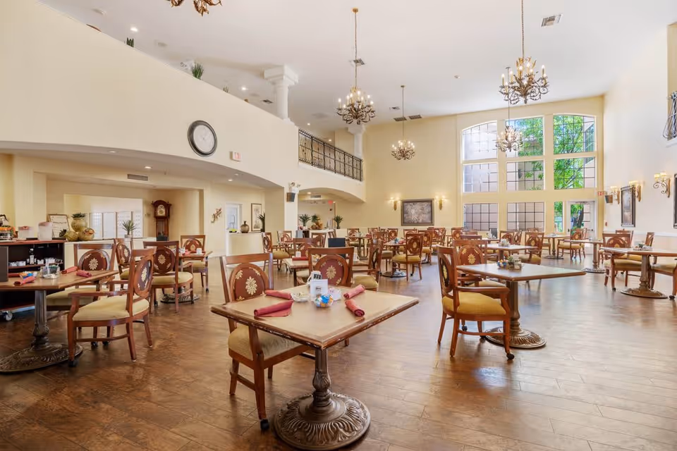 A spacious dining room in a senior living facility with multiple wooden tables and chairs arranged neatly. The room features high ceilings with chandeliers, large windows allowing natural light to fill the space, and a clock mounted on a cream-colored wall. The floor is wooden, and the tables are set with red napkins and small decorative items.