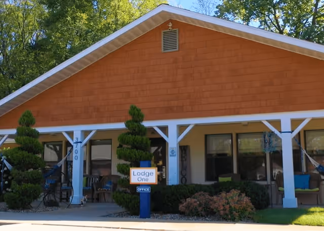 Exterior view of a single-story building with a brown shingled roof and white pillars supporting a covered porch. The porch has chairs and hammocks, with neatly trimmed bushes and small trees in front. A sign near the entrance reads 'Lodge One OFFICE'.