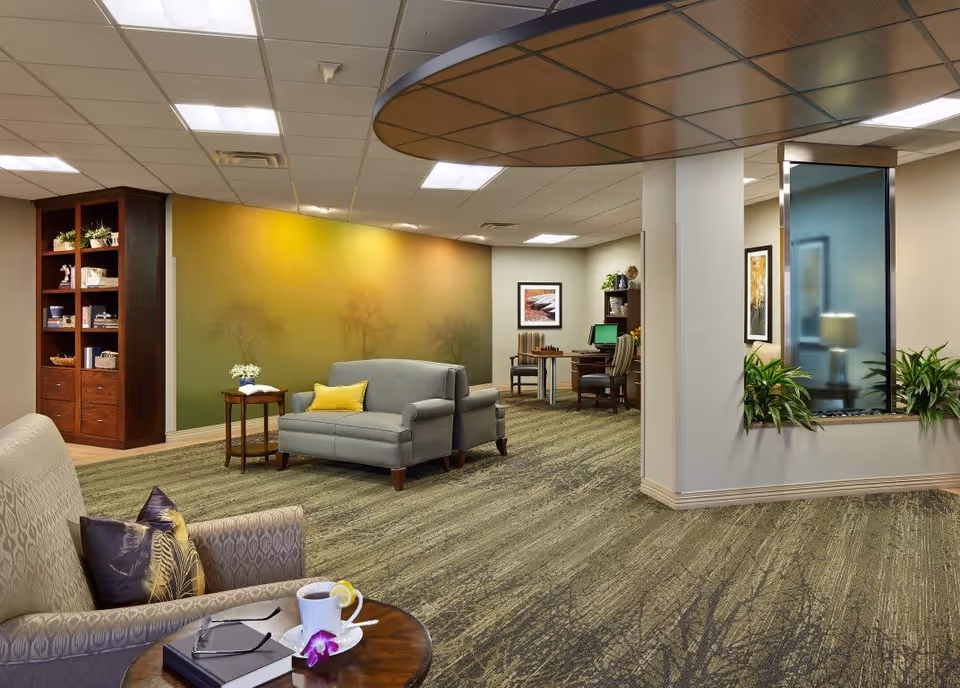 A cozy and well-lit living room area in a senior living facility with comfortable armchairs and a loveseat, a small wooden side table with a cup of tea and a book, a green and yellow gradient wall with subtle tree designs, a wooden bookshelf with plants and decorative items, and a small office area with a desk, computer, and chairs in the background.