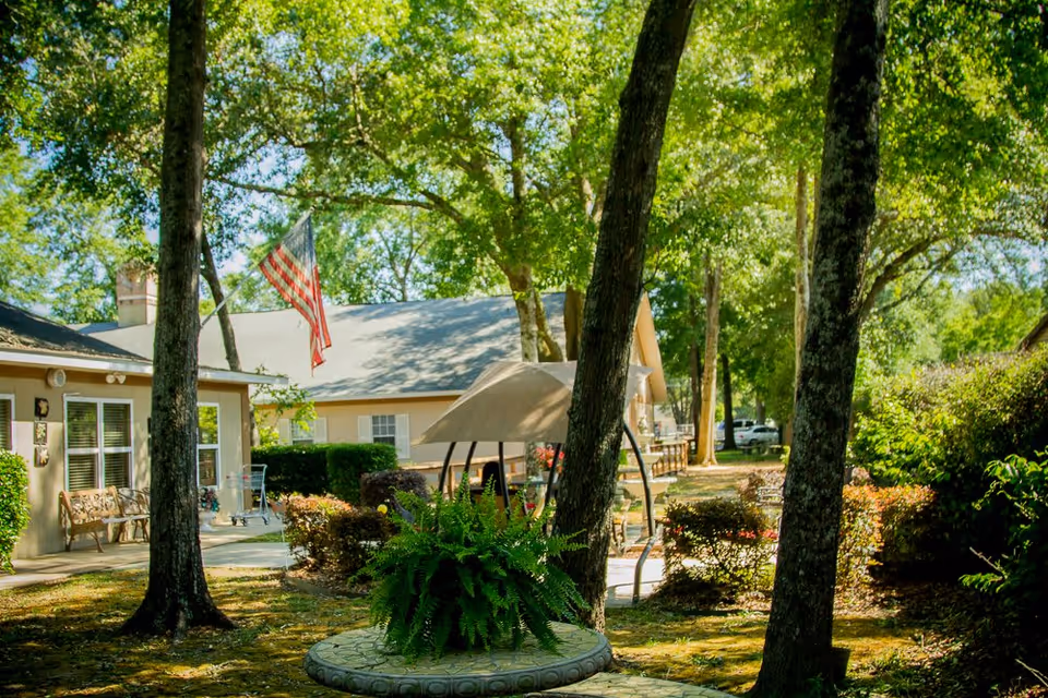 Shaded courtyard with trees, a stone planter holding a fern, outdoor seating and an American flag in front of a single-story building.