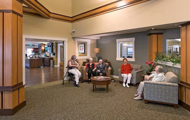 A group of elderly people sitting and chatting in a spacious, well-lit senior living facility common area with comfortable chairs, a coffee table, and decorative plants. The room has carpeted flooring and wooden trim, with an adjacent kitchen or dining area visible in the background.
