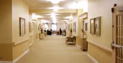 A long, well-lit hallway in a senior living facility with beige walls and carpeted floor. The hallway is decorated with framed pictures on the walls, small tables, and chairs along the sides. At the far end, there is a seating area with people gathered around tables.