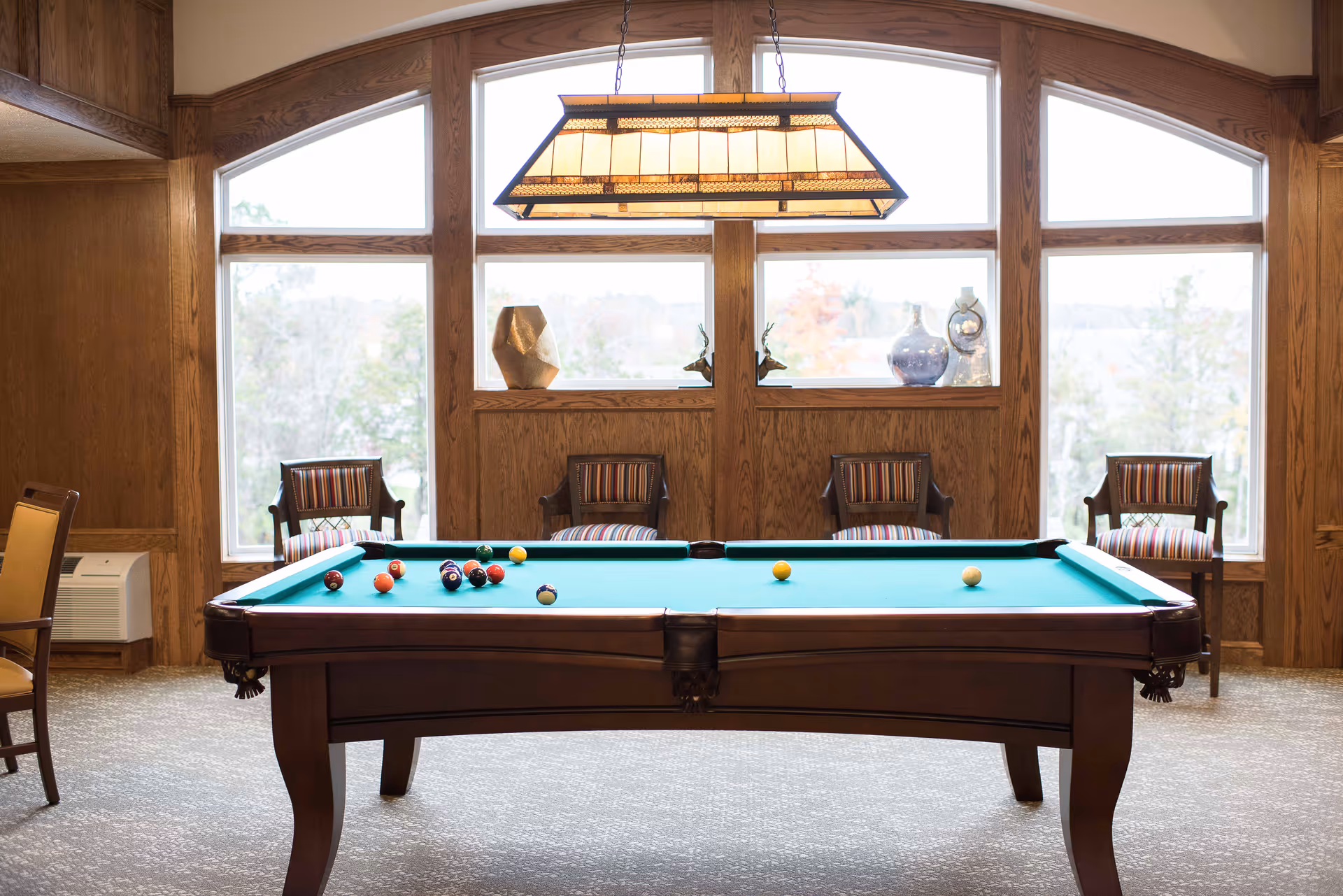 A pool table with billiard balls arranged on the green felt surface in a room with large windows and wooden paneling. Four striped cushioned chairs are placed against the wall under the windows, and a rectangular hanging light fixture is above the pool table.