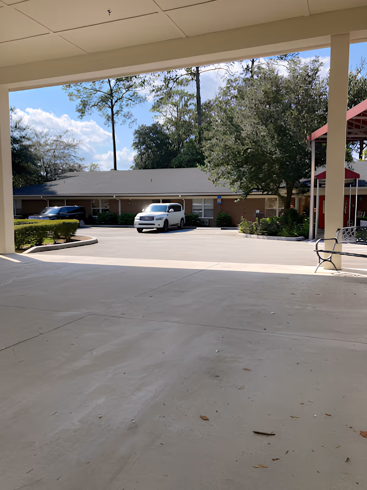 View from a covered outdoor area looking out onto a parking lot with two parked vehicles, surrounded by trees and bushes under a partly cloudy sky.