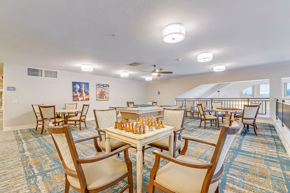 A bright and spacious senior living common area with multiple tables and chairs arranged for social activities. In the foreground, there is a chess table with chess pieces set up. In the background, there is a pool table and additional seating. The room has modern ceiling lights and a ceiling fan, with large windows allowing natural light to enter.